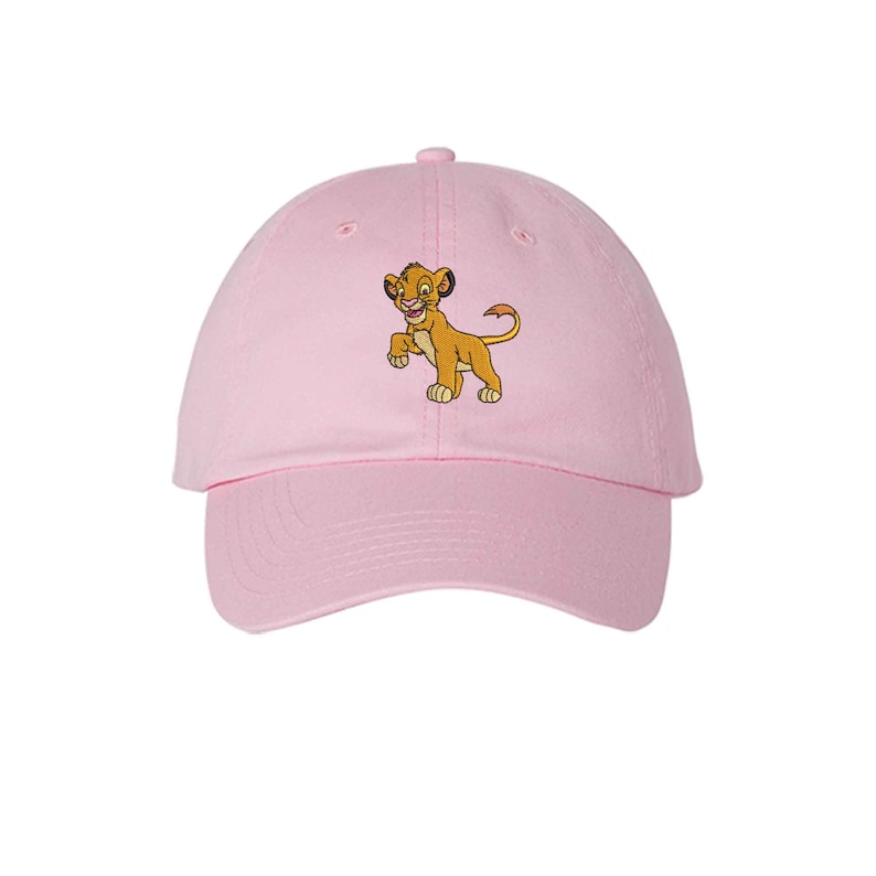 May include: A light pink baseball cap featuring an embroidered graphic of a young lion cub with golden-brown fur, dark ear tips, and a tufted tail. The cap has a curved brim, a button on the top, and stitched eyelets for ventilation.