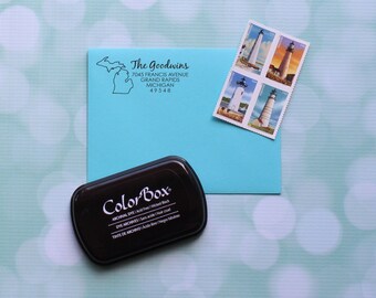 May include: A blue envelope with a return address that reads "The Goodwins, 7045 Francis Avenue, Grand Rapids, Michigan 49548". The envelope has a sheet of four lighthouse stamps attached to the upper right corner. A black ink pad with the label "ColorBox" is in the lower left corner of the image.