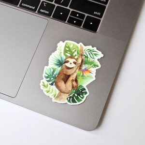 May include: A watercolor illustration of a sloth hanging from a tree branch surrounded by tropical leaves and flowers. The sloth is brown with a white face and black eyes. The leaves are green and the flowers are orange, yellow, and purple.