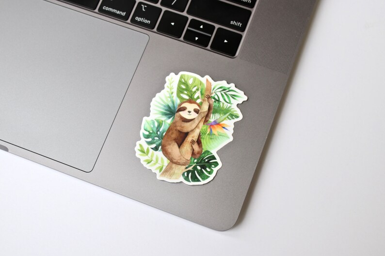 May include: A watercolor illustration of a sloth hanging from a tree branch surrounded by tropical leaves and flowers. The sloth is brown with a white face and black eyes. The leaves are green and the flowers are orange, yellow, and purple.