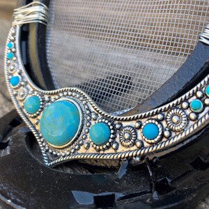 May include: A silver horseshoe with a turquoise and silver design. The horseshoe is decorated with a row of turquoise stones and silver beads. The horseshoe is on a black background.