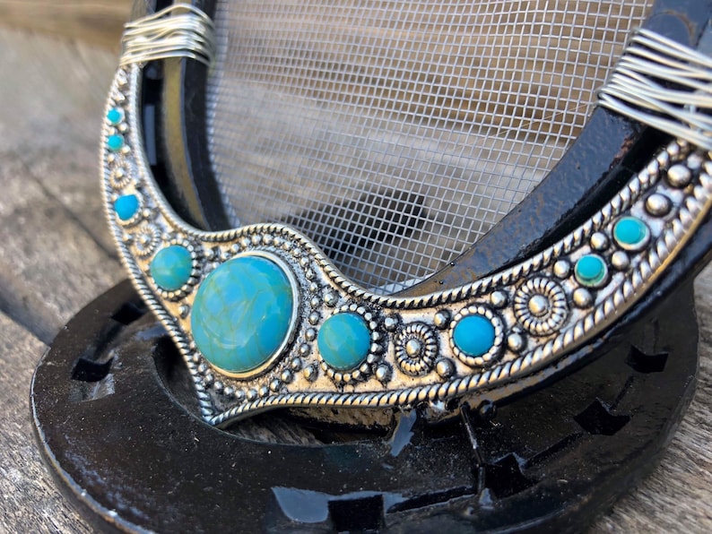 May include: A silver horseshoe with a turquoise and silver design. The horseshoe is decorated with a row of turquoise stones and silver beads. The horseshoe is on a black background.
