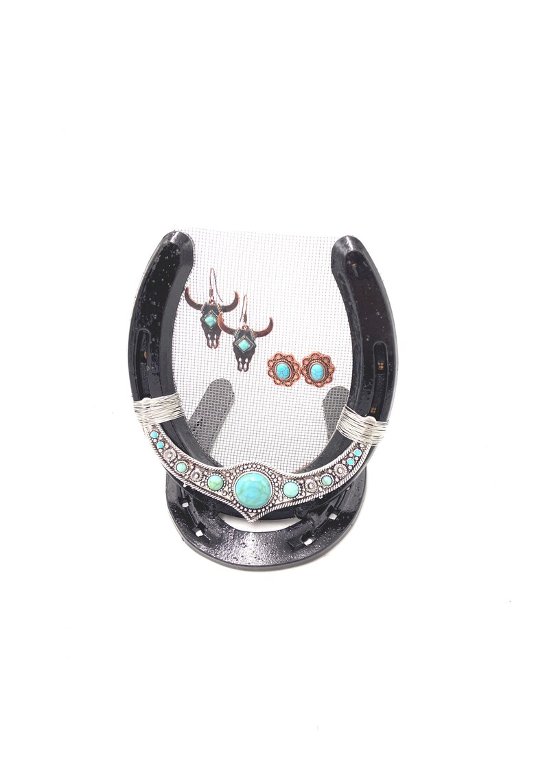 May include: A black horseshoe with a wire mesh center, decorated with turquoise and silver accents, used as a jewelry stand.  The stand is displaying a pair of turquoise and silver earrings and a pair of turquoise and copper earrings.