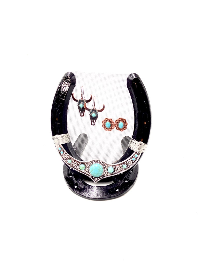 May include: A black horseshoe-shaped jewelry stand with turquoise and silver accents. The stand is decorated with a turquoise and silver design and features a mesh backing. The stand is displaying a pair of turquoise and silver earrings and a pair of turquoise and silver stud earrings.