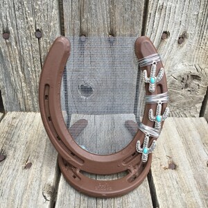 Pu&ograve; includere: Un ferro di cavallo marrone con un retro in rete metallica e tre ciondoli a forma di cactus argentati con pietre turchesi. Il ferro di cavallo &egrave; su una superficie di legno.