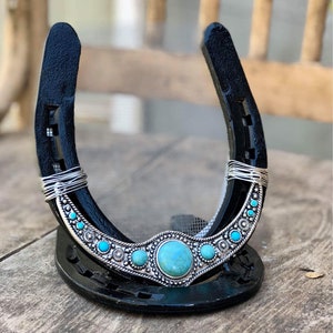 May include: A black horseshoe with a silver and turquoise design. The horseshoe is decorated with a large turquoise stone in the center and smaller turquoise stones on either side. The horseshoe is standing upright on a wooden surface.