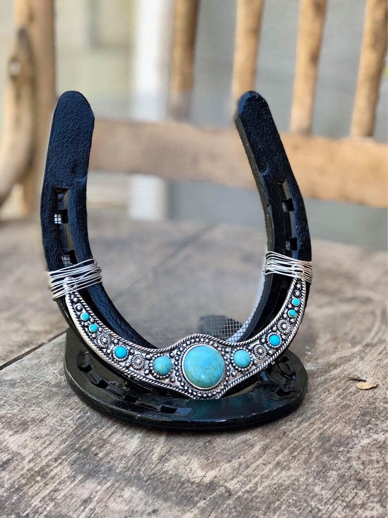 May include: A black horseshoe with a silver and turquoise design. The horseshoe is decorated with a large turquoise stone in the center and smaller turquoise stones on either side. The horseshoe is standing upright on a wooden surface.