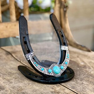 May include: A black horseshoe with a turquoise and silver design. The horseshoe is decorated with a large turquoise stone in the center and smaller turquoise stones around the edge. The horseshoe is on a wooden surface.