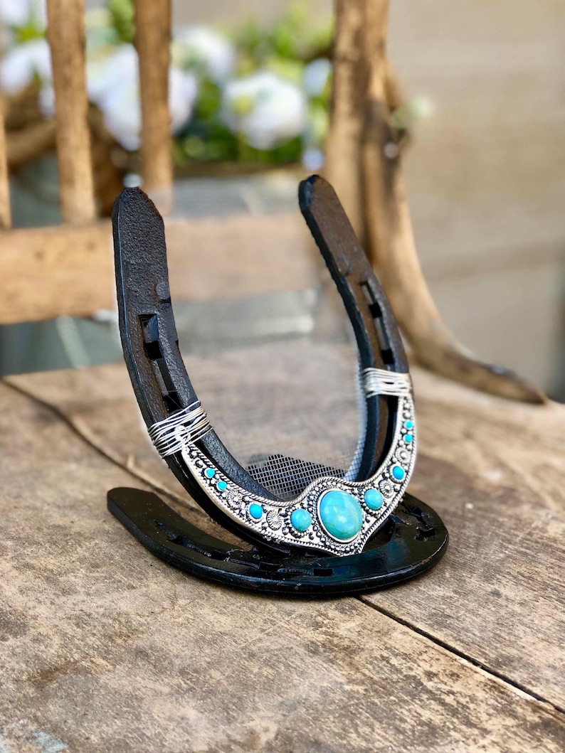May include: A black horseshoe with a turquoise and silver design. The horseshoe is decorated with a large turquoise stone in the center and smaller turquoise stones around the edge. The horseshoe is on a wooden surface.