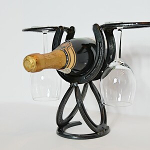 May include: Black metal wine bottle holder with two glasses hanging from the side. The bottle holder is made of horseshoes.