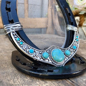 May include: A black horseshoe with a wire-wrapped turquoise and silver design. The horseshoe is decorated with turquoise stones and silver accents. The horseshoe is a decorative object.