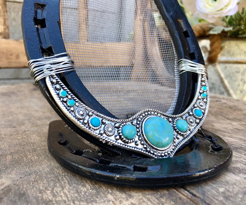 May include: A black horseshoe with a wire-wrapped turquoise and silver design. The horseshoe is decorated with turquoise stones and silver accents. The horseshoe is a decorative object.