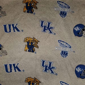 May include: Gray fabric with a repeating pattern of blue and yellow Kentucky Wildcats logos, basketball hoops, and football helmets. The logos include the letters "UK" and the word "Wildcats".