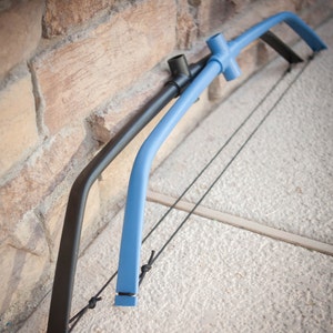 May include: A blue and black PVC pipe bow with a string attached. The bow is leaning against a brick wall.