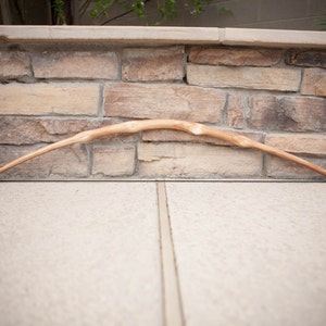 May include: A wooden bow with a natural finish, resting on a stone wall. The bow is curved and has a slight bend in the middle.