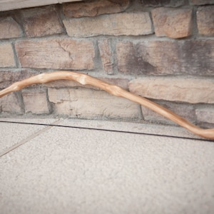 May include: A wooden bow with a natural finish and a black string. The bow is resting on a concrete surface.