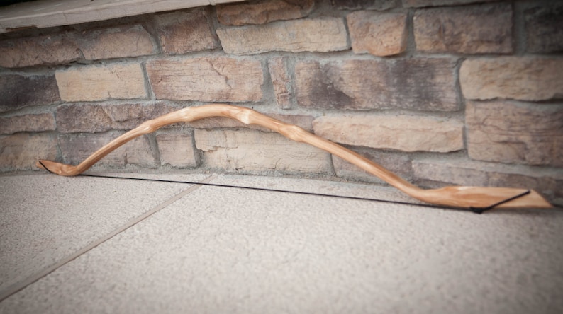 May include: A wooden bow with a natural finish and a black string. The bow is resting on a concrete surface.