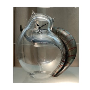 May include: A clear glass pitcher with an owl-like design. The pitcher has a curved handle with red and grey stripes. The clock hands are visible on the front. The pitcher is on a white surface.