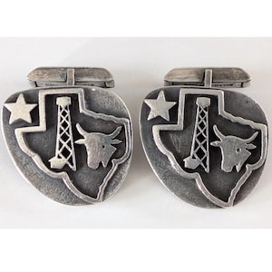 May include: A pair of silver-coloured cufflinks. Each cufflink features a Texas-shaped design with a star, oil derrick, and longhorn. The cufflinks have a textured, antique finish and a hinged closure.