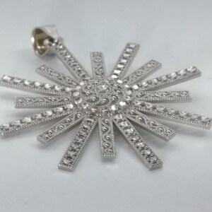 May include: A silver-toned pendant in a sunburst design. The pendant features a central cluster of small, faceted stones surrounded by radiating rays, each adorned with smaller stones and decorative detailing. The bail is a simple loop.