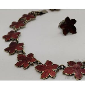 Puede incluir: Un conjunto de joyas vintage que incluye un collar y un anillo. El collar está compuesto por colgantes rojos en forma de flor con detalles metálicos dorados. El anillo tiene un diseño de flor roja a juego. El conjunto se exhibe sobre una superficie blanca.