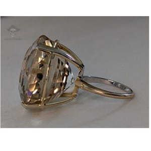 May include: A ring featuring a large, faceted, champagne-colored gemstone set in a gold-toned metal prong setting. The band is thin and metallic, showing a two-tone appearance with a gold-colored top section and a silver-colored lower portion.