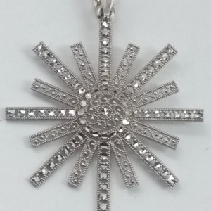 May include: A silver-toned pendant in a sunburst design. The pendant features a central circular element with intricate detailing, surrounded by radiating rectangular bars. Each bar is adorned with small, sparkling accents, creating a dazzling effect. The pendant has a loop for a chain.