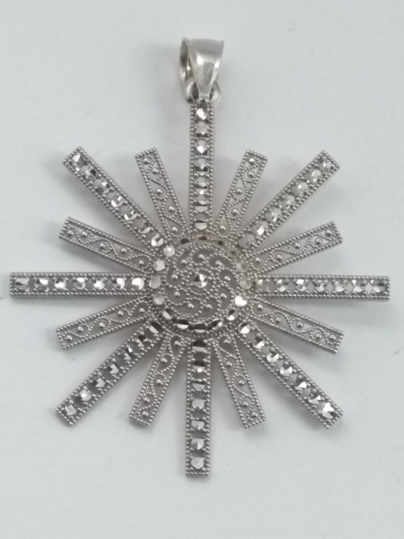 May include: A silver-toned pendant in a sunburst design. The pendant features a central circular element with intricate detailing, surrounded by radiating rectangular bars. Each bar is adorned with small, sparkling accents, creating a dazzling effect. The pendant has a loop for a chain.