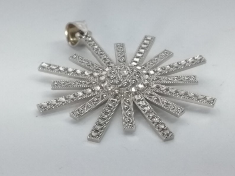 May include: A silver-toned pendant in a sunburst design. The pendant features a central cluster of small, faceted stones surrounded by radiating rays, each adorned with smaller stones and decorative detailing. The bail is a simple loop.