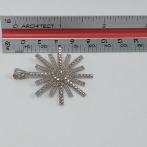 May include: A silver-toned pendant in a sunburst design, adorned with small, sparkling stones. The pendant is approximately 1.5 inches in diameter. The pendant has a loop for a chain and is set against a white background.