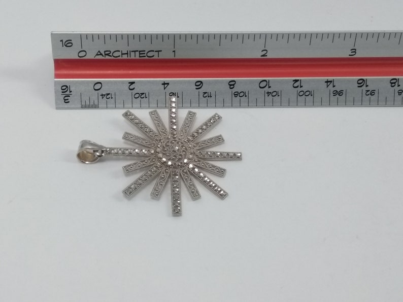 May include: A silver-toned pendant in a sunburst design, adorned with small, sparkling stones. The pendant is approximately 1.5 inches in diameter. The pendant has a loop for a chain and is set against a white background.