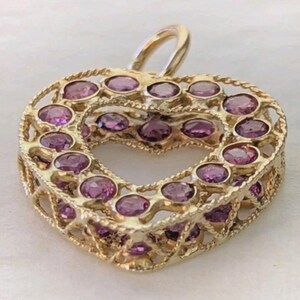May include: A heart-shaped pendant crafted from gold-toned metal, featuring a lattice-like frame adorned with numerous round, faceted purple gemstones set throughout the front and side surfaces, complete with a small gold-toned bail at the top.