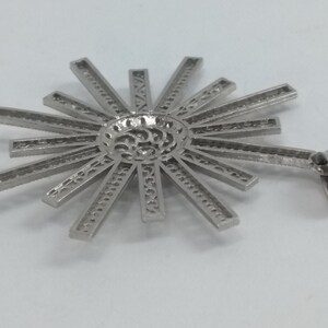 May include: A silver-colored pendant with a sunburst design. The pendant features a central circular element with intricate cutouts, from which radiate numerous rectangular rays. A small loop is attached for hanging. The pendant is likely made of metal.