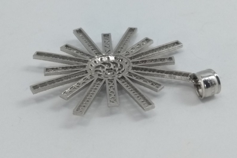 May include: A silver-colored pendant with a sunburst design. The pendant features a central circular element with intricate cutouts, from which radiate numerous rectangular rays. A small loop is attached for hanging. The pendant is likely made of metal.