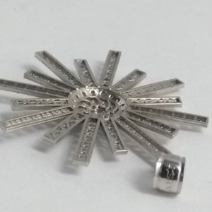 May include: A silver-colored pendant with a sunburst design. The pendant features a central, textured circle with radiating rectangular bars. A small, cylindrical loop is attached for a chain.