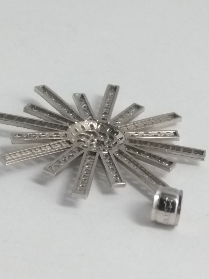 May include: A silver-colored pendant with a sunburst design. The pendant features a central, textured circle with radiating rectangular bars. A small, cylindrical loop is attached for a chain.