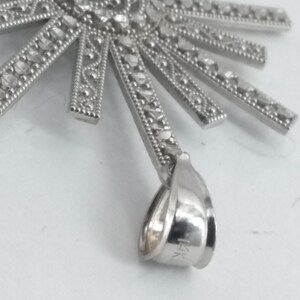 May include: A silver-toned pendant with a sunburst design. The pendant features a central circular element with intricate detailing and radiating rectangular rays, each adorned with small, sparkling accents. The bail is marked with "14K".