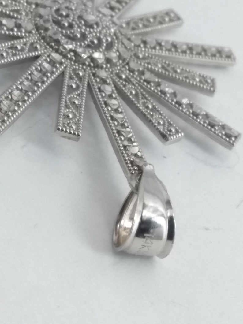 May include: A silver-toned pendant with a sunburst design. The pendant features a central circular element with intricate detailing and radiating rectangular rays, each adorned with small, sparkling accents. The bail is marked with "14K".
