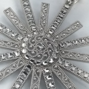 May include: A silver-toned pendant with a sunburst design. The pendant features a central spiral and radiating beams, each adorned with clear, sparkling stones. The beams are accented with intricate detailing, creating a dazzling effect. The pendant has a loop for a chain.