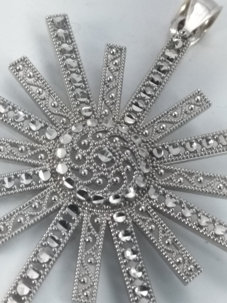 May include: A silver-toned pendant with a sunburst design. The pendant features a central spiral and radiating beams, each adorned with clear, sparkling stones. The beams are accented with intricate detailing, creating a dazzling effect. The pendant has a loop for a chain.