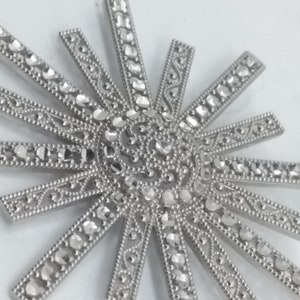 May include: Close-up of a silver-toned sunburst pendant. The pendant features a central circular design with radiating rays, each adorned with small, sparkling accents. The intricate details and bright finish create a striking visual.