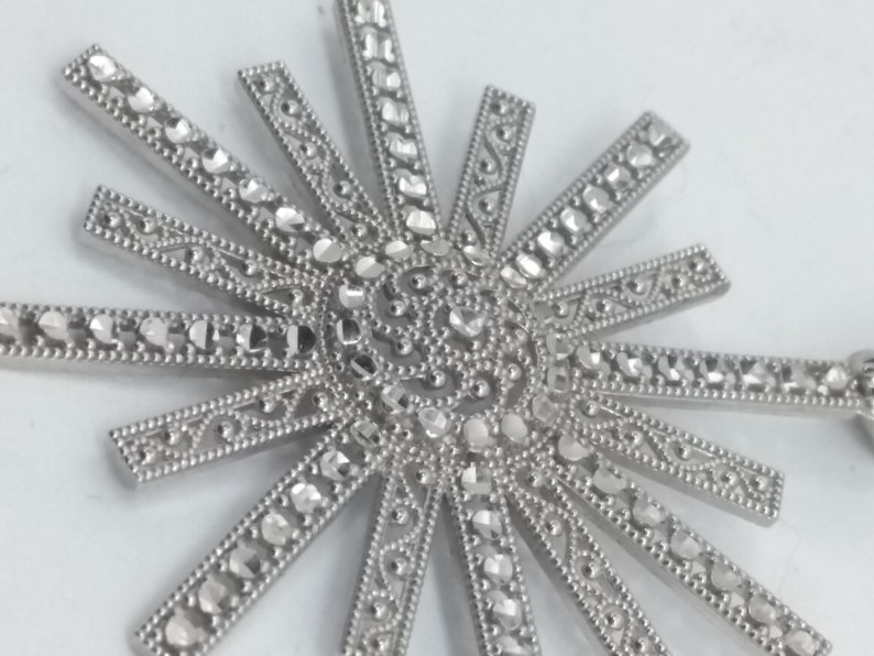 May include: Close-up of a silver-toned sunburst pendant. The pendant features a central circular design with radiating rays, each adorned with small, sparkling accents. The intricate details and bright finish create a striking visual.