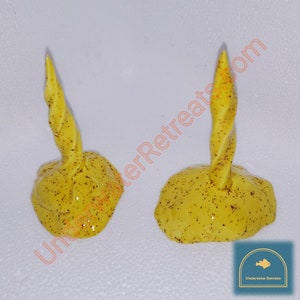May include: Two yellow, abstract candle holders with a textured, speckled design. Each has a rounded base and a twisted, upward-pointing element. The candle holders are decorated with small, dark brown speckles.