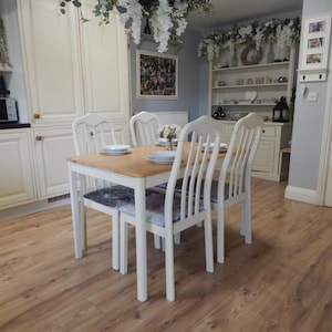 May include: A dining table and four white chairs with grey upholstered seats. The table has a light wood top. The room features light-coloured cabinets and a microwave. The floor is wood laminate.