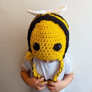 May include: A crocheted bee hat with black and yellow stripes, white wings, and black eyes. The hat is being worn by a child.