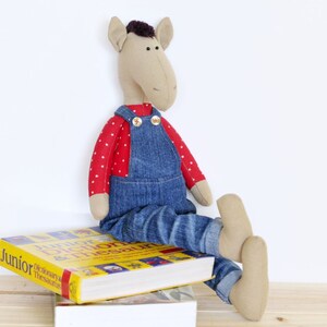 Op de afbeelding: Een bruin knuffelpaardje dat een rood-wit geblokt shirt en een blauwe denim overall draagt. Het paardje zit op een geel en rood boek met de titel "Junior Dictionary & Thesaurus".