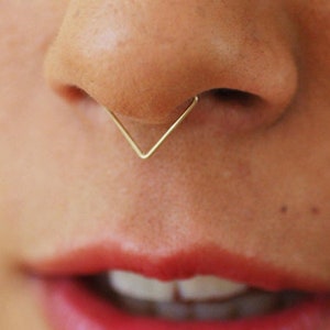 Könnte beinhalten: Ein goldener Nasenring in Form eines Dreiecks. Der Ring wird im Septum-Piercing getragen.