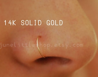 Może przedstawiać: Zbliżenie na nos osoby z małym, cienkim złotym kółkiem do nosa. Tekst "14K SOLID GOLD" znajduje się nad kółkiem do nosa, a tekst "junelittlehop.etsy.com" pod kółkiem do nosa.