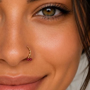 Peut inclure: Un anneau de nez en or avec un design de chaîne perlée délicate et deux petites perles rondes en pierre précieuse rose foncé suspendues en bas. Le bijou est porté dans la narine, avec une finition dorée métallique et des accents roses.
