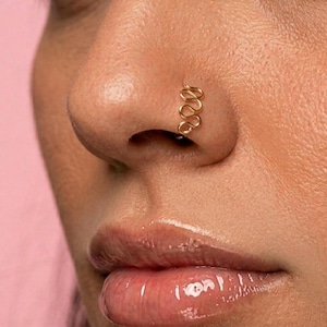 May include: A close-up of a nose with a gold-colored nose ring. The nose ring has a looped design. The skin is a light tone, and the background is a soft pink.